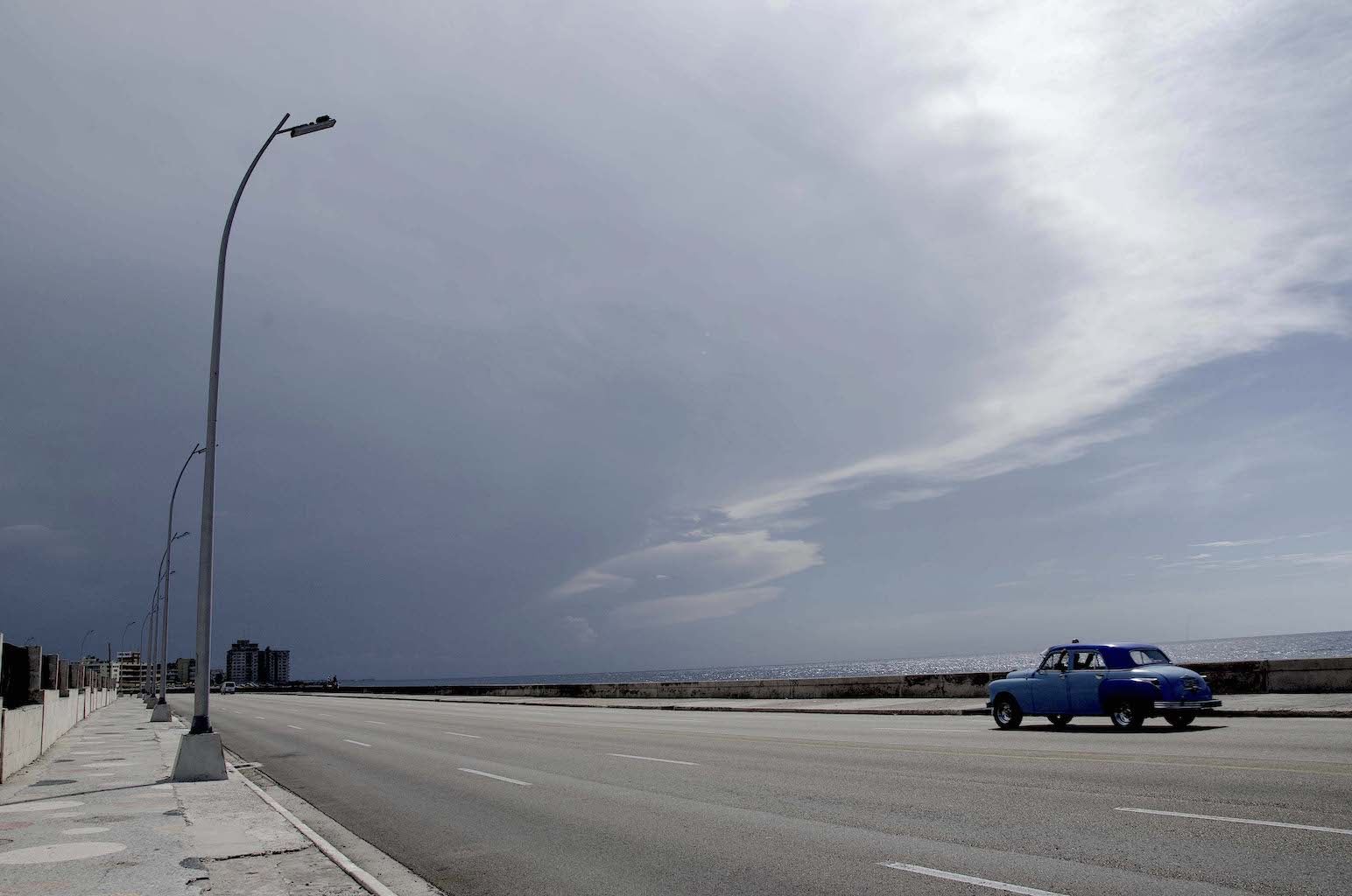 Photo of a blue car beneath a cloudy sky along the Malecón Seawall in Havana, Cuba.